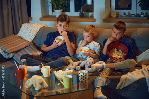 Children, brothers and little sister sitting on couch in living room in evening, watching tv together and eating snacks. Concept of family, leisure time, relaxation, childhood and parenthood
