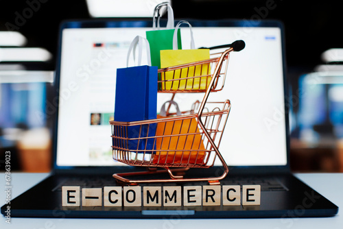 E-commerce and online shopping concept. Shopping cart, laptop, colourful bags in the office. Blurred background.