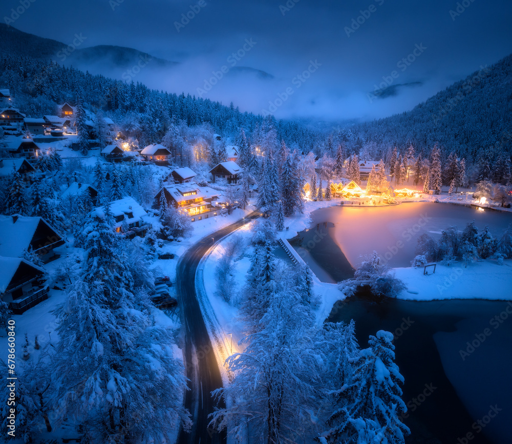 Aerial view of fairy town in snow, road, forest, Jasna lake and houses with lights at night in winter. Top view of mountain village, illumination, snowy pine trees at dusk in Kranjska Gora, Slovenia - obrazy, fototapety, plakaty 