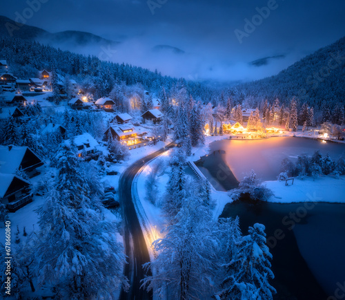 Aerial view of fairy town in snow, road, forest, Jasna lake and houses with lights at night in winter. Top view of mountain village, illumination, snowy pine trees at dusk in Kranjska Gora, Slovenia