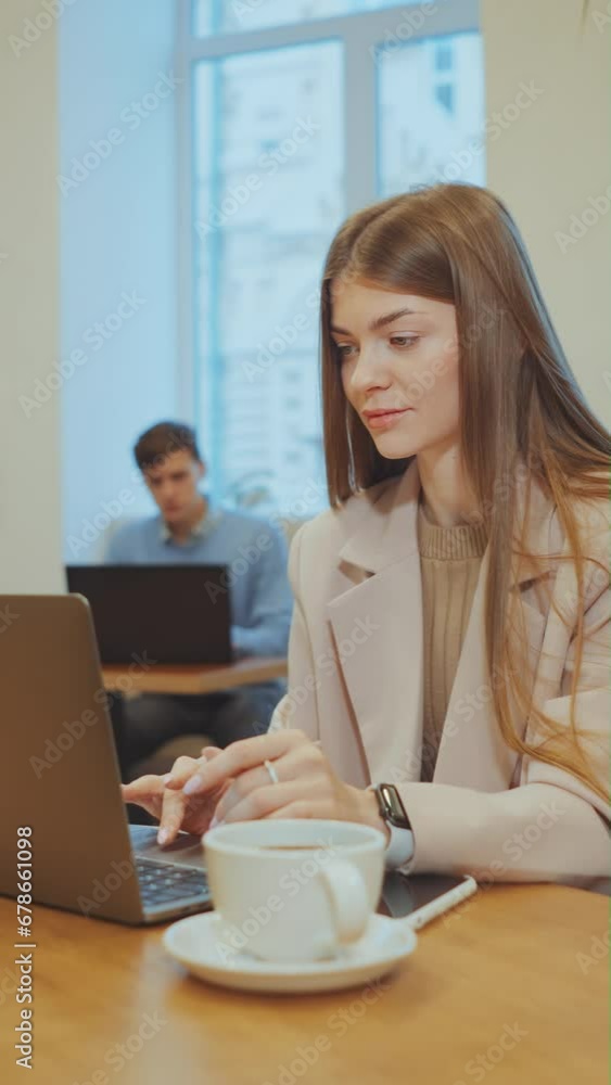Vertical Screen: Woman wearing jacket typing on laptop in cafe, coffee cup and glass of water standing beside her on table, man using laptop by window on blurred background. Concept of freelance Vertical Screen: Woman wearing jacket typing on laptop in cafe, coffee cup and glass of water standing beside her on table, man using laptop by window on blurred background. Concept of freelance