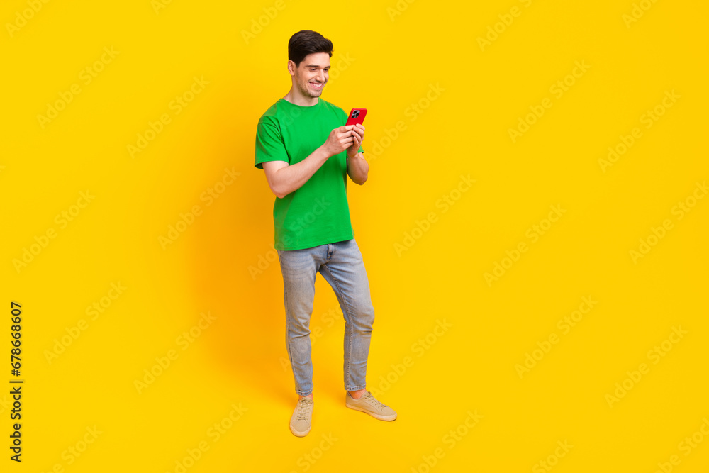 Full size photo of good mood guy wear stylish t-shirt denim pants look at smartphone read notification isolated on yellow color background Full size photo of good mood guy wear stylish t-shirt denim pants look at smartphone read notification isolated on yellow color background
