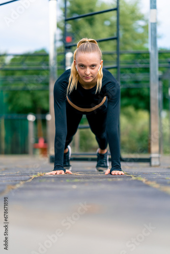 On a sports street playground a pumped-up girl stands in plank and listens to motivating music on headphones