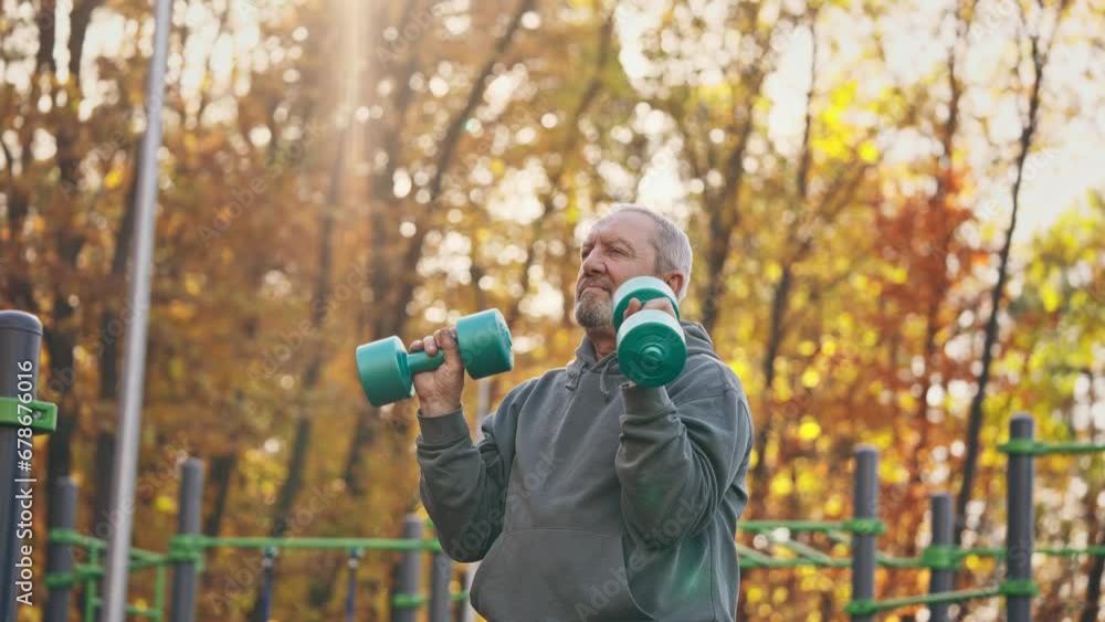 An active grey-haired elderly man in his retirement age doing exercises with dumbbells on his own outdoors. Grandpa does sports An active grey-haired elderly man in his retirement age doing exercises with dumbbells on his own outdoors. Grandpa does sports