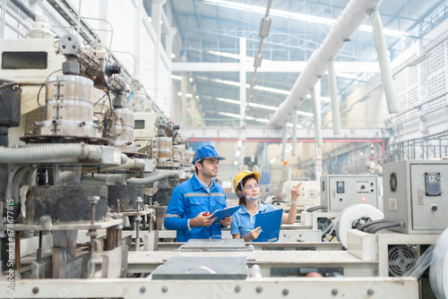 American male engineer and Asian female manager point to destination Wear a uniform and safety helmet. There are machines working in a factory, industrial business producing plastics and steel.