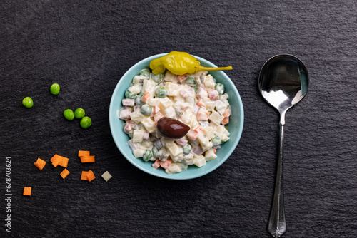 A bowl with the appetizer Ruska Salata, Bulgarian variation of Olivier salad, placed on black stone next to a spoon, spread green peas, diced pickled carrots  and garnished with an olive and pepper © Ivelin