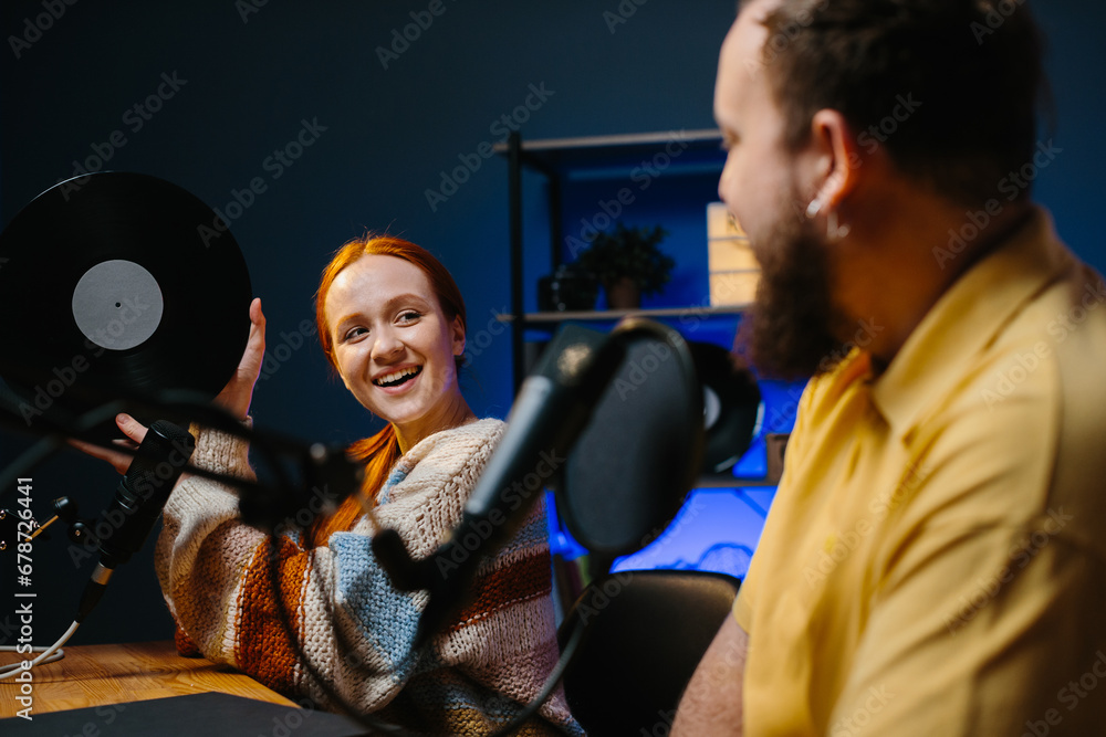 Music bloggers recording a new episode of their podcast. A man and a woman are talking about music in the studio, the woman is holding a vinyl record in her hands.