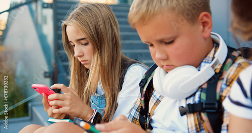 Close up of group of schoolchildren sitting on stairs outdoors using mobile phone communicating online. Diverse kids use internet on cellphone while relaxing on street at sunny day