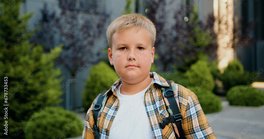 Close up of pensive good- looking caucasian schoolboy with backpack posing and looking at camera in school building background, outdoors. Elementary preteen school student portrait