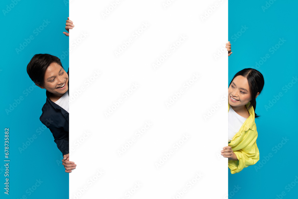 Curious smiling young korean man and lady in casual look at big banner with copy space for words Curious smiling young korean man and lady in casual look at big banner with copy space for words