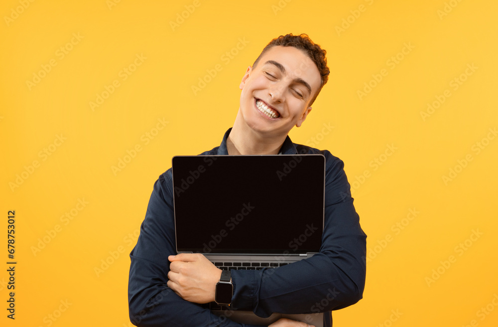 Joyful young guy hugging open laptop, yellow background