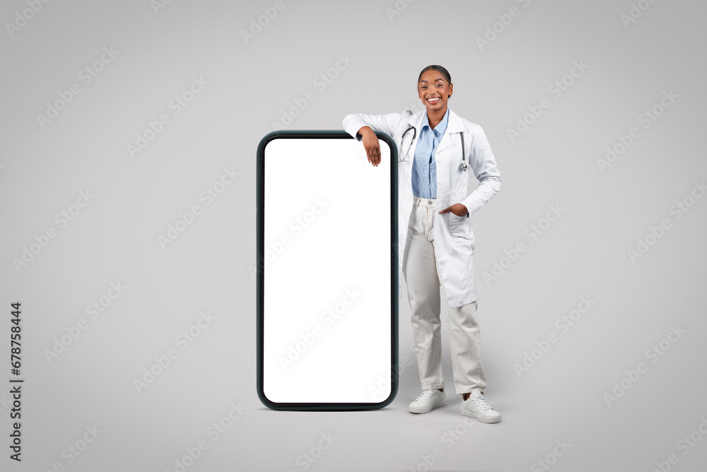 Happy confident black millennial doctor in white coat near big mobile smartphone with blank screen Happy confident black millennial doctor in white coat near big mobile smartphone with blank screen