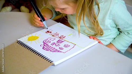 Child draws a family next to a house in a sketchbook with colored markers.