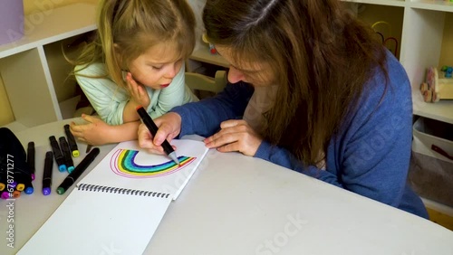 Child with mom draws rainbow in sketchbook on table with colored markers.