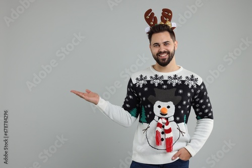 Young man in Christmas sweater and reindeer headband showing something on grey background. Space for text photo