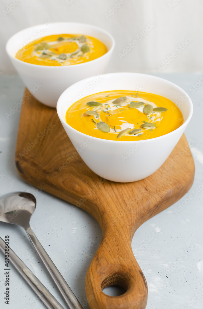 Two white bowls of pumpkin cream soup with dried seeds and thyme leaves on a wooden cutting board on a gray background. Concept of healthy eating. Vegetarian and vegan food. Vertical orientation. Two white bowls of pumpkin cream soup with dried seeds and thyme leaves on a wooden cutting board on a gray background. Concept of healthy eating. Vegetarian and vegan food. Vertical orientation.