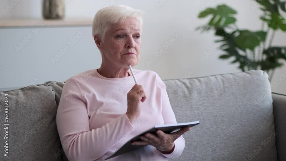 Dreamy eyed Caucasian elderly woman sits on the couch, face illuminated by the spark of creativity. With a sense of purpose, she scribbles ideas into a notebook. Dreamy eyed Caucasian elderly woman sits on the couch, face illuminated by the spark of creativity. With a sense of purpose, she scribbles ideas into a notebook.