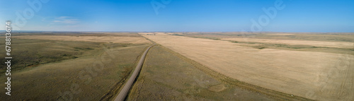Aerial panoramic view of an extensive dry prairie landscape under a bright blue sky. A rough looking road crosses the fields and disappears into the horizon. 