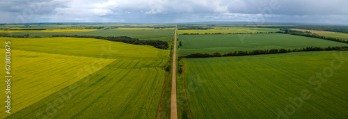 Aerial panoramic view of a vast prairie agricultural landscape with wheat and canola fields and a long straight gravel farm road. The road disappears into the distance. The sky is full of gray clouds 