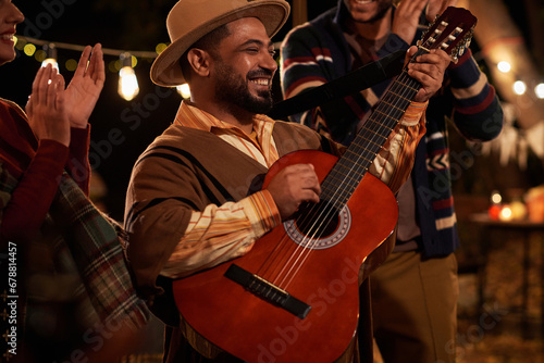 Happy man in traditional clothes playing guitar at holiday outdoors with other people dancing