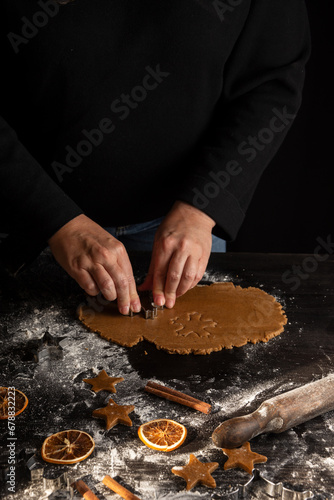 Top view of woman's hands with mold on gingerbread cookie dough on dark wooden table with cinnamon, flour, oranges and rolling pin, vertical