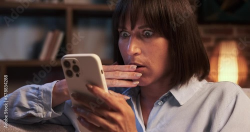 Startled Caucasian woman clutches phone tightly in the living room, eyes wide with fear. The room seems to echo her sense of unease, the atmosphere tense and apprehensive. photo