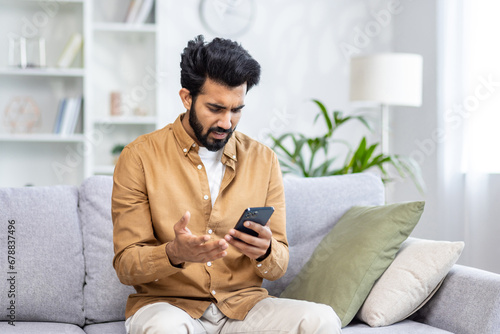 Sad unhappy and disappointed man sitting at home on sofa in living room, Indian man received notification message with bad news, reading using smartphone app.