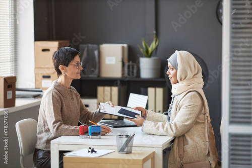 Side view of mature manager of visa application center and young Muslim female applicant wearing hijab and quiet luxury attire
