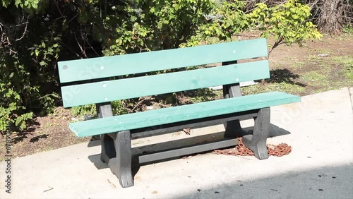 empty green gray solo single wood wooden bench park outside on pavement cement with tree bush blowing behind it photo