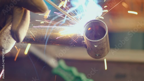 Hand in protective glove welding metal pipe details together with welding torch producing flame, fume and sparks. Close-up view, focus of foreground photo