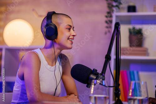 A cheerful gen z girl is sitting in a small recording studio at home and wearing headphones while speaking at the microphone during the podcast.