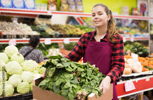 Portrait of a positive young saleswoman near the counter in a store, holding a box of spinach