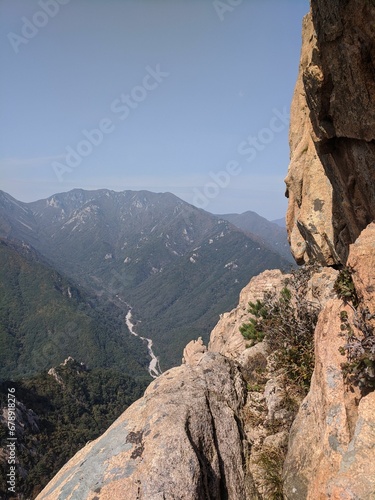 Vertical view from a steep cliff to a mountain range, dense forests and a river on a sunny day © Wirestock