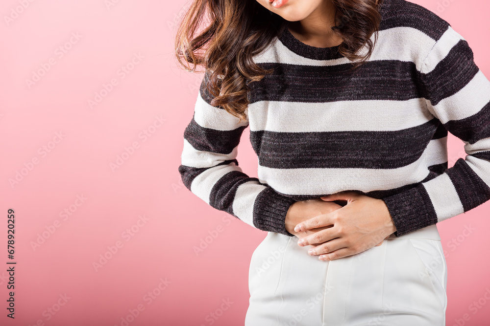 An Asian woman's portrait shows her with stomachache and abdominal suffering in a studio shot against a pink background. Depicting health and medical issues, specifically gastritis. An Asian woman's portrait shows her with stomachache and abdominal suffering in a studio shot against a pink background. Depicting health and medical issues, specifically gastritis.