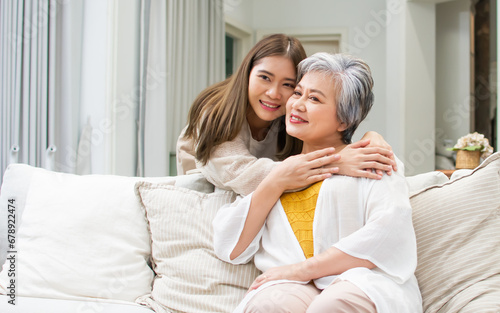 Happy Asian adorable sweet beautiful adult daughter hugging her mother with happiness, love, warmth, smiling, sitting on sofa at cozy home. Family, Healthcare, Insurance, Retirement Concept.