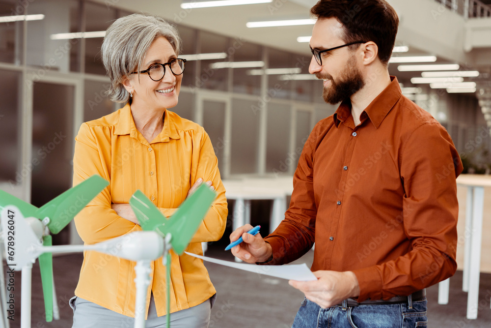 Obraz premium Team of smiling business partners engineers, developers talking, planning startup project, cooperation using windmill working in modern office. Alternative, wind energy, innovation concept