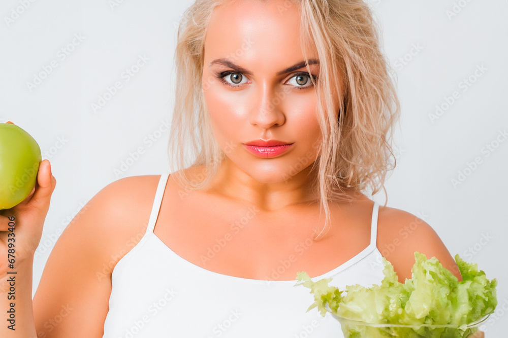 A beautiful blonde woman holds a green apple in her right hand and a transparent glass bowl of lettuce in her left hand A beautiful blonde woman holds a green apple in her right hand and a transparent glass bowl of lettuce in her left hand