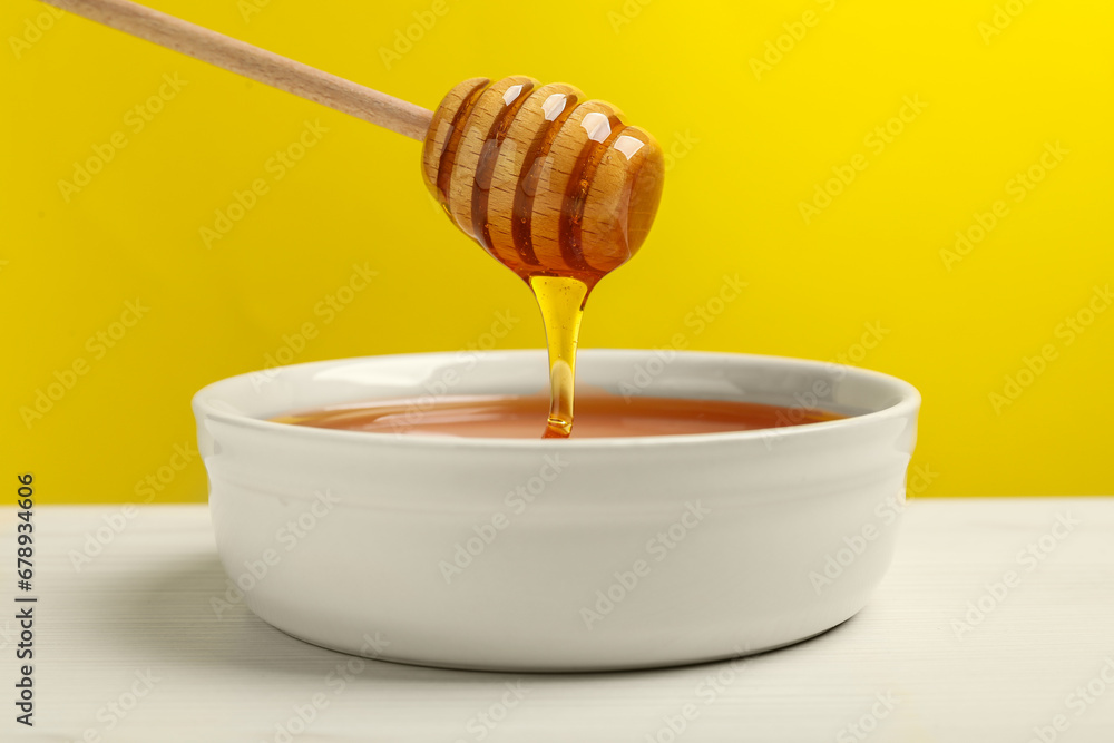 Pouring tasty honey from dipper into bowl at white wooden table against yellow background, closeup Pouring tasty honey from dipper into bowl at white wooden table against yellow background, closeup