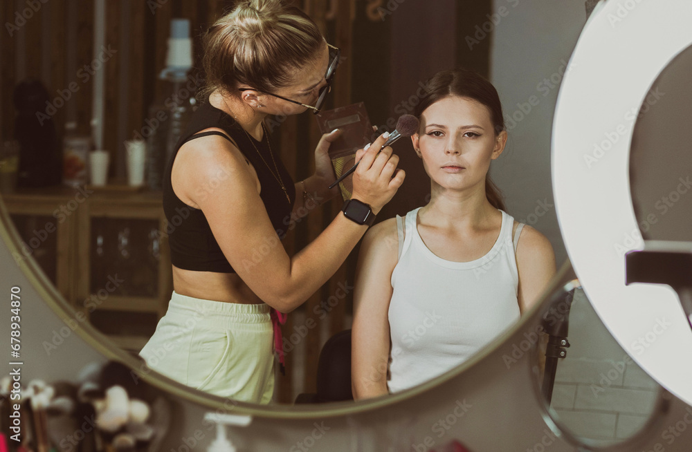 A young makeup artist applies foundation powder to a girl face with a brush. A young makeup artist applies foundation powder to a girl face with a brush.