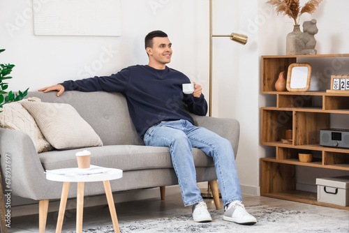 Handsome young man with cup of coffee sitting on sofa in modern living room © Pixel-Shot