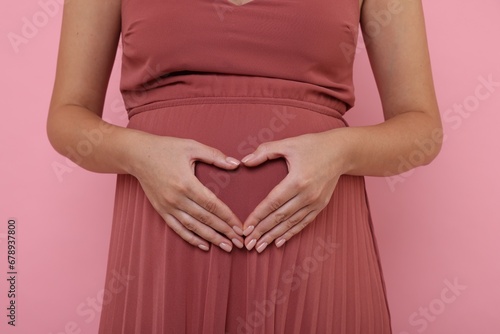 Pregnant woman in dress making heart with hands on pink background, closeup