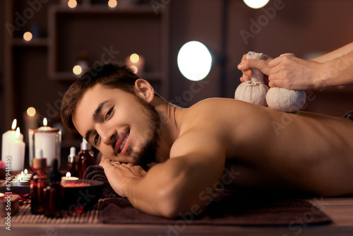 Young man undergoing treatment with herbal bags in dark spa salon, closeup