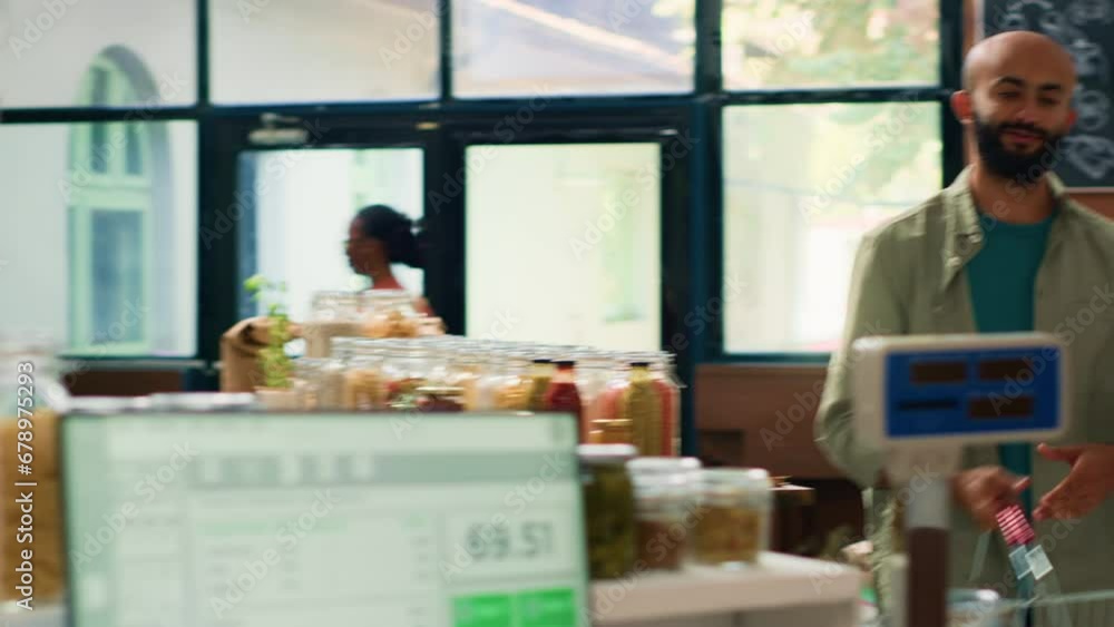 Salesperson in convenience store weighs fresh produce and groceries for middle eastern shopper by using digital scale at register. Retailer in local shop selling bulk pantry goods. Handheld shot. Salesperson in convenience store weighs fresh produce and groceries for middle eastern shopper by using digital scale at register. Retailer in local shop selling bulk pantry goods. Handheld shot.