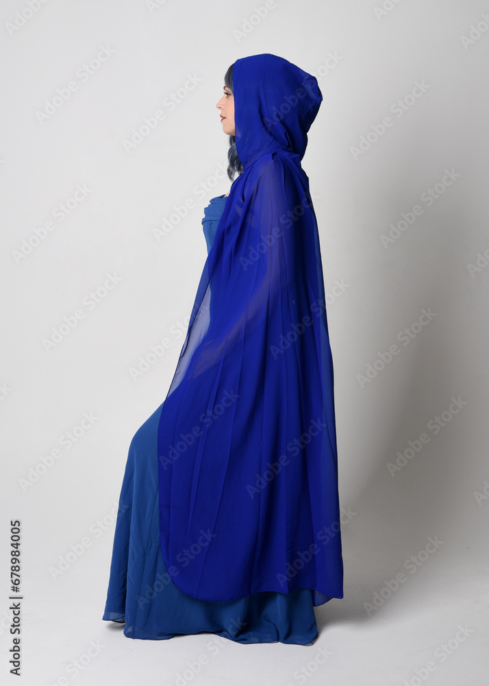 Full length portrait of beautiful young female model elegant blue fantasy  ball gown and flowing cape with hood. Standing pose, walking away facing backwards. Isolated on whit studio background.