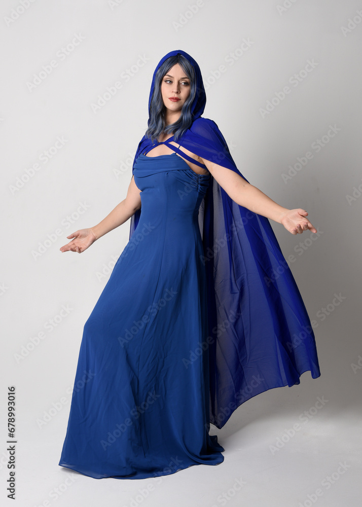 Full length portrait of beautiful female model wearing elegant fantasy blue ball gown and flowing cape with hood.
Standing pose, with gestural arms reaching out . Isolated on white studio background.