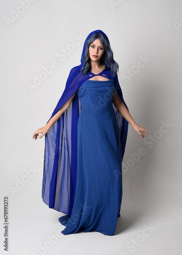 Full length portrait of beautiful female model wearing elegant fantasy blue ball gown and flowing cape with hood. Standing pose, with gestural arms reaching out . Isolated on white studio background.