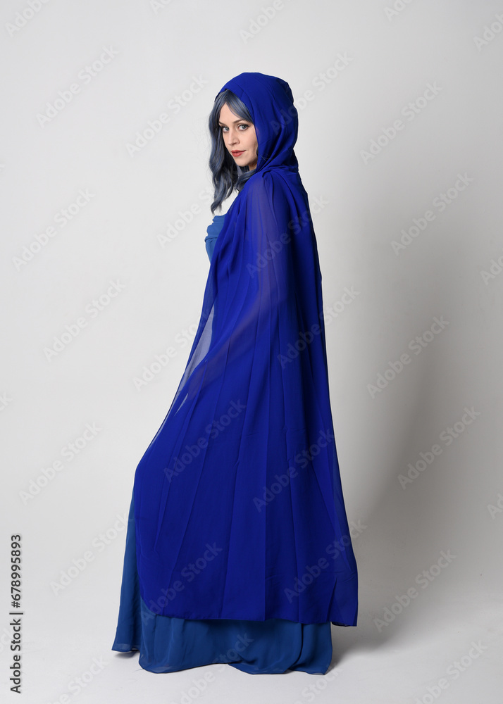 Full length portrait of beautiful female model wearing elegant fantasy blue ball gown, flowing cape with hood.
Standing pose walking away, gestural arms reaching out. Isolated on studio background Full length portrait of beautiful female model wearing elegant fantasy blue ball gown, flowing cape with hood.
Standing pose walking away, gestural arms reaching out. Isolated on studio background