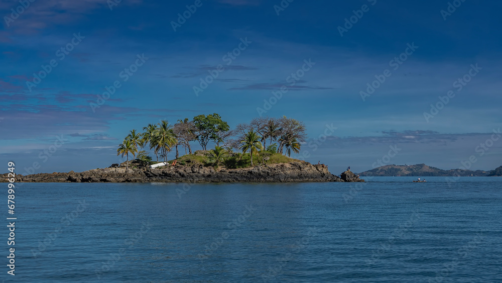 Naklejka premium A beautiful tropical island in the blue ocean. Palm trees grow on the rocks. Tiny silhouettes of people and boats are visible. A mountain range in the distance against the azure sky and clouds.