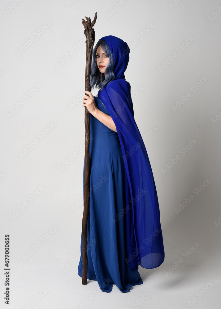Full length portrait of beautiful female model wearing elegant fantasy blue ball gown, flowing cape with hood.Standing pose walking away, holding a wooden wizard staff. Isolated on white studio backgr Full length portrait of beautiful female model wearing elegant fantasy blue ball gown, flowing cape with hood.Standing pose walking away, holding a wooden wizard staff. Isolated on white studio backgr