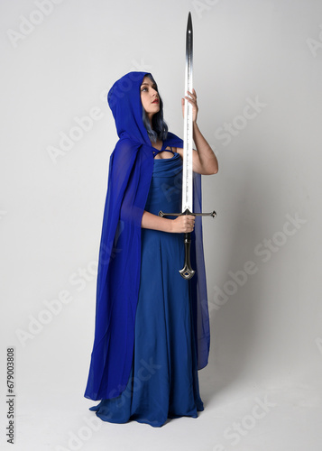 Full length portrait of beautiful female model wearing elegant fantasy blue ball gown, flowing cape with hood. Standing pose walking away, holding a sword weapon Isolated on white studio background.